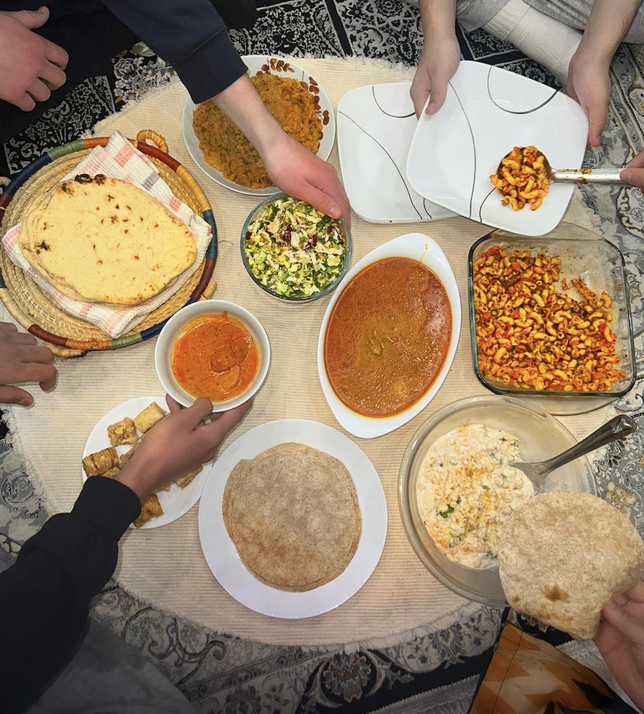 A bird’s eye view of several hands reaching for food, such as butter chicken, macaroni salad, roti and baklava.