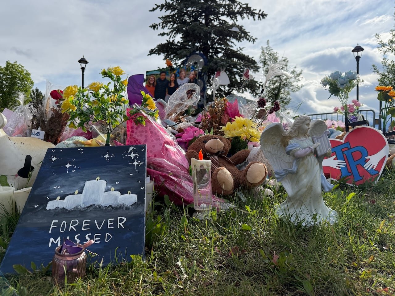 default-6 outdoor memorial in front of a spruce tree with flowers, candles, teddy bears, photos of children, and an angel statue beside a sign that reads “FOREVER MISSED”